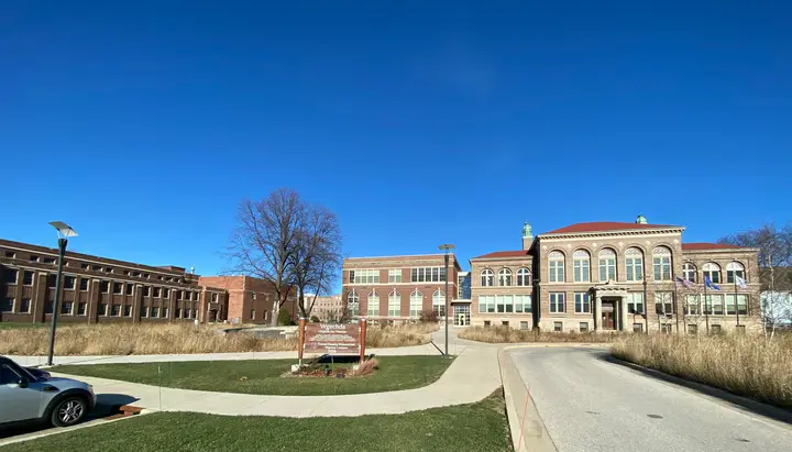 A picture of the campus today on a clear, winter day. There is a long 3 story brick building to the left; another complex of two 3 story buildings starting in the middle. In front are fields.