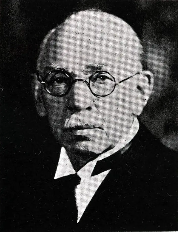 Black & white portrait of an elderly man wearing a suit and tie. He wears round eye-glasses and is bald on top.