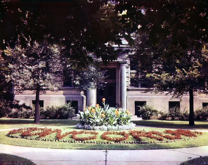 The same building in summer blocked by trees with a garden out front. The garden, surrounded by a walkway contains flowers that spell out 