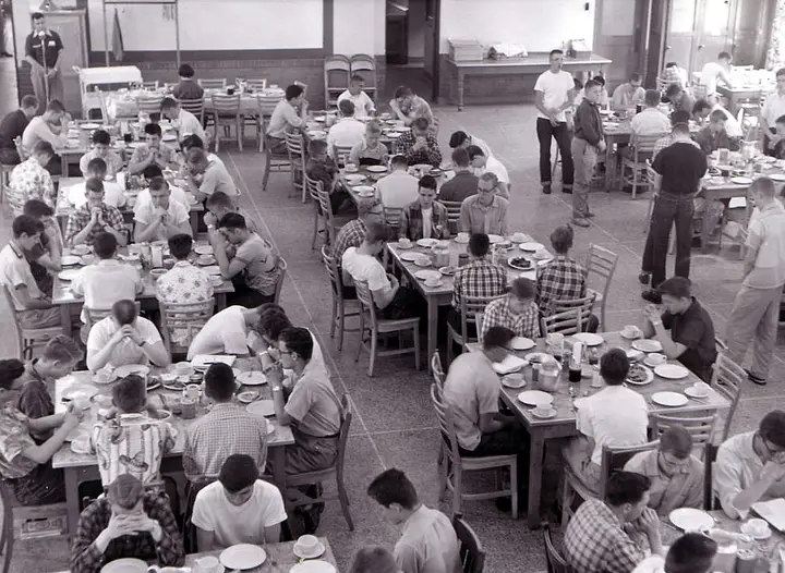 Black and white photo of a cafeteria where many young men are praying before their meal.