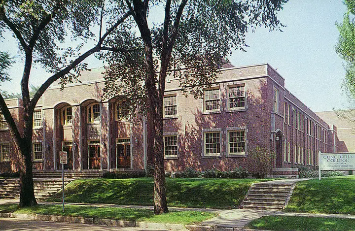 2.5 story brick gymnasium; shaded by trees on a sunny day. Picture is taken from an angle.