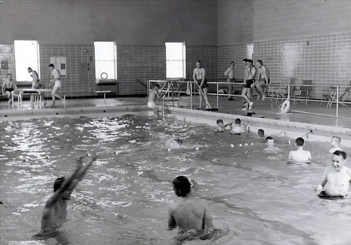 Black and white photo of multiple people relaxing in or on the side of an indoor swimming pool.