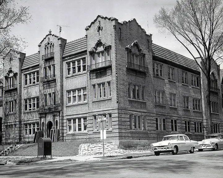 Black and white photo of a 3 story brick building with 50's style cars parked alongside.
