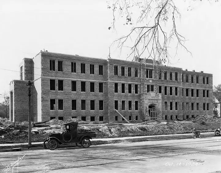 Black and white photo of a 3.5 story brick building under construction. An old 20's style car is parked out front with a motorcyle behind it.