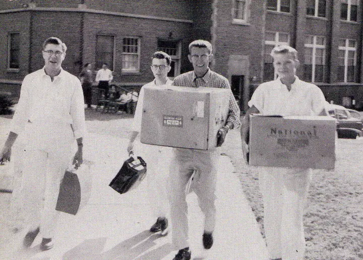 4 men in a black and white photo carrying boxes on the campus.