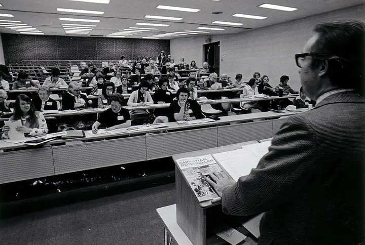 Mostly female students receiving a lecture from a man at a lectern at the front of the classroom.