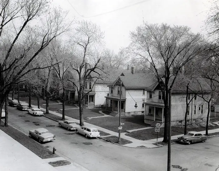 Black and white photo of a block of 2.5 story houses with cars parked in front.