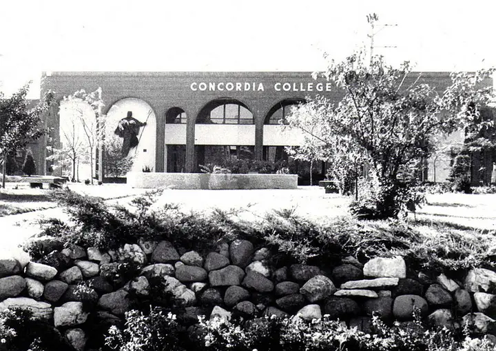 2 story brick college building with a large green space in front. A small stone retaining wall is in the forefront.